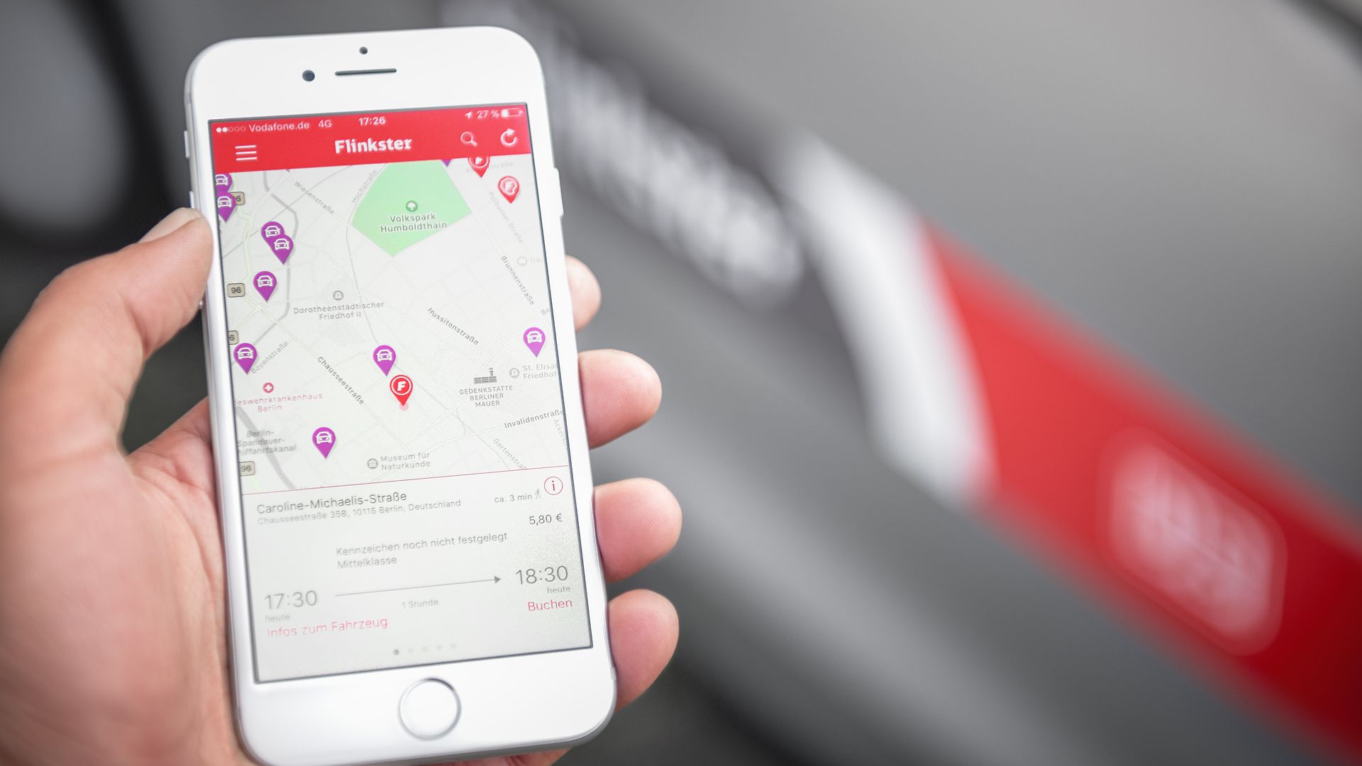 One hand holds a smartphone with the Flinkster app open. A vehicle with the DB logo can be seen blurred in the background.