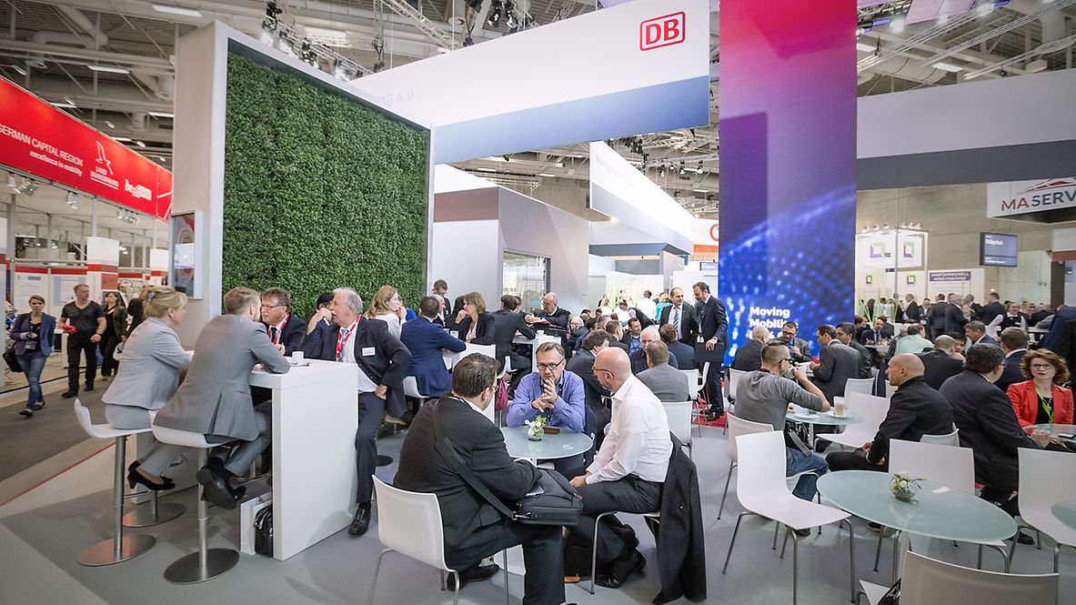 Deutsche Bahn exhibition booth with a wall of plants, the DB logo and lots of people sitting at tables and chatting.