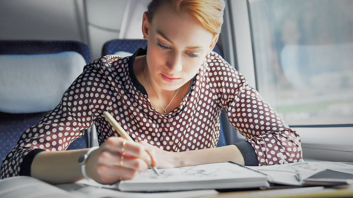 A business traveler sits by the window on an ICE train and writes something in a notebook.