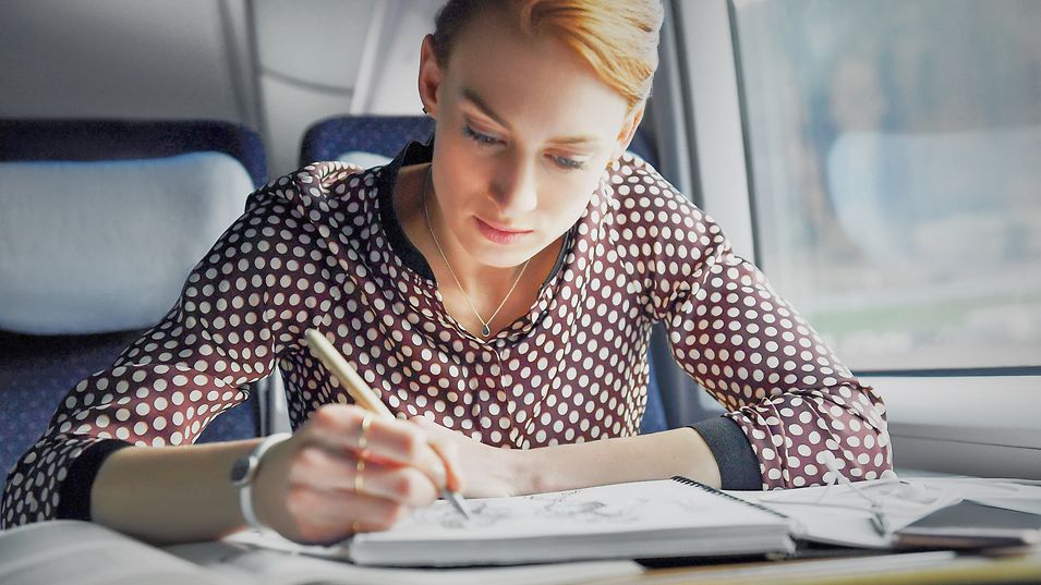 A business traveler sits by the window on an ICE train and writes something in a notebook.