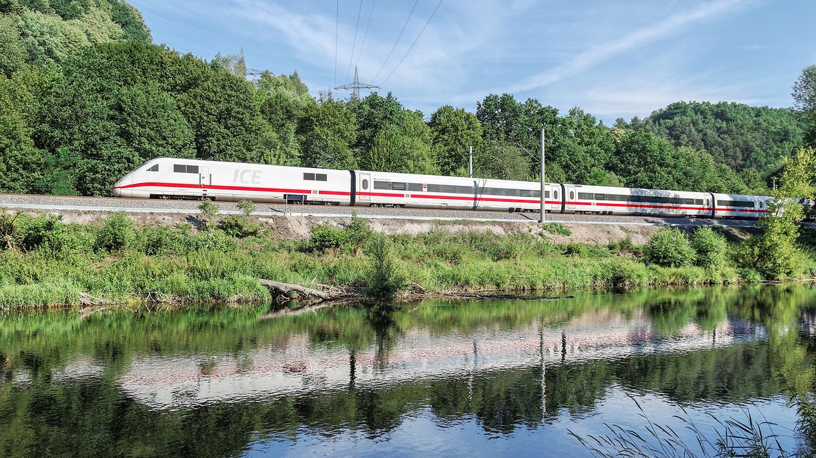 An ICE train travels along a river through green countryside.
