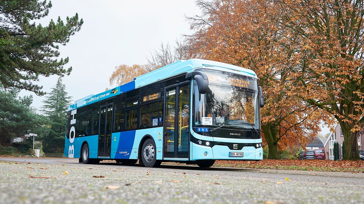A light blue bus stands on a road with autumnal trees in the background