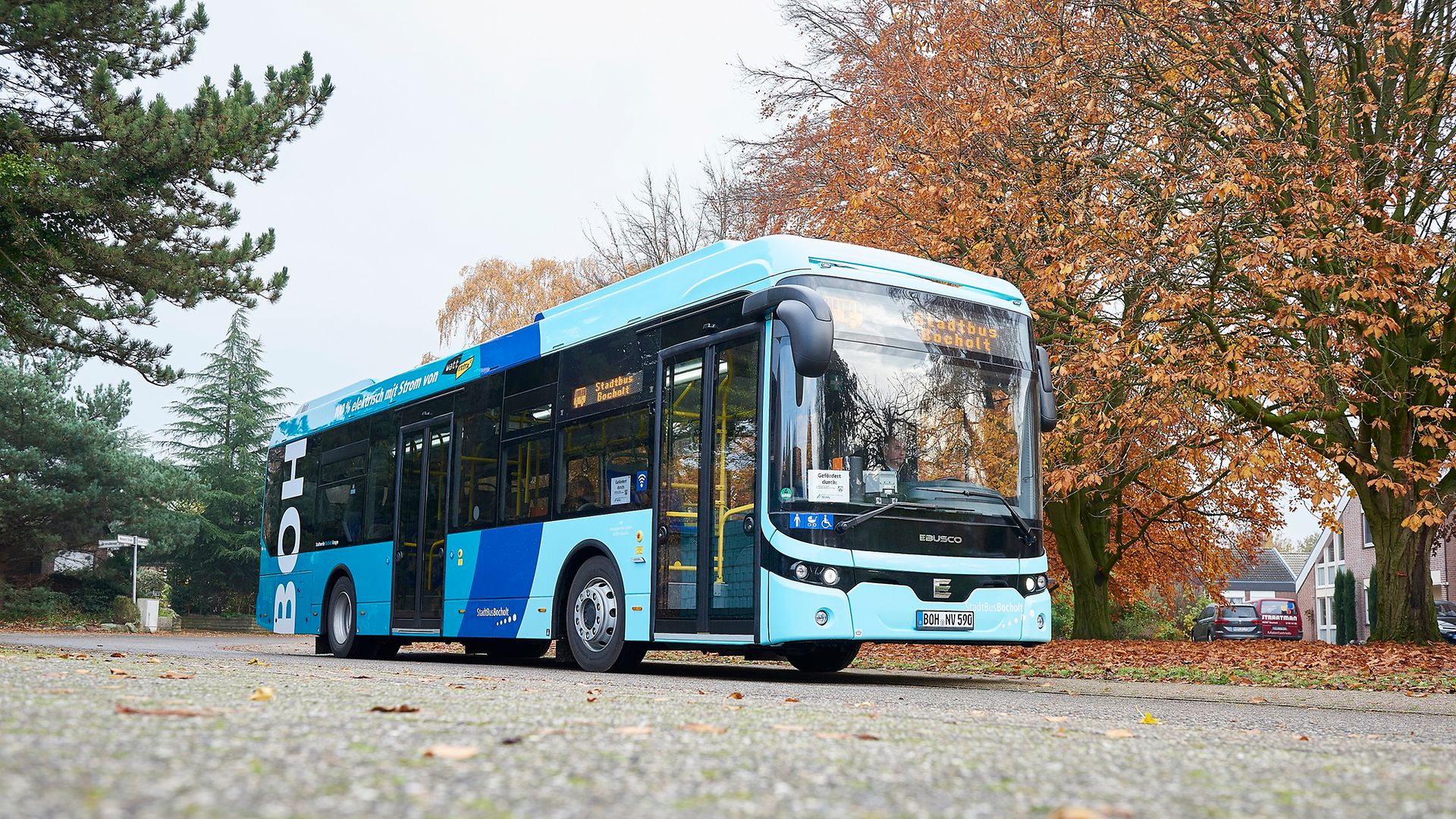 A light blue bus stands on a road with autumnal trees in the background