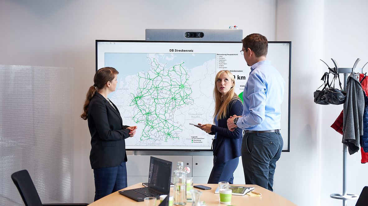 Three people are standing in a meeting room in front of a screen showing a map of Germany.