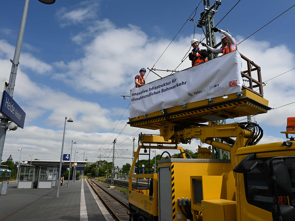 Three construction workers are standing on the lifting platform of a rail construction vehicle and working on the overhead line.