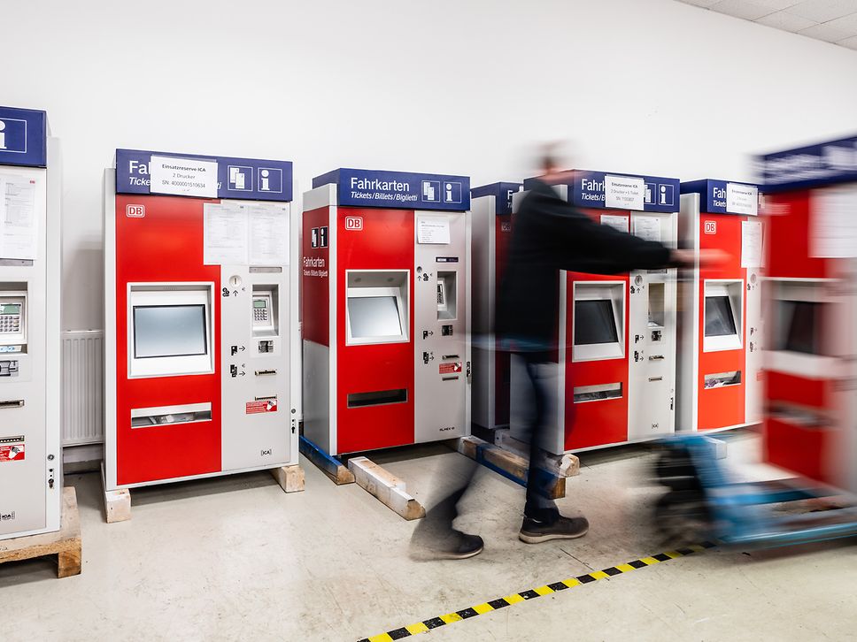 Some ticket machines stand in a depot. A man with a lift truck walks past. 
