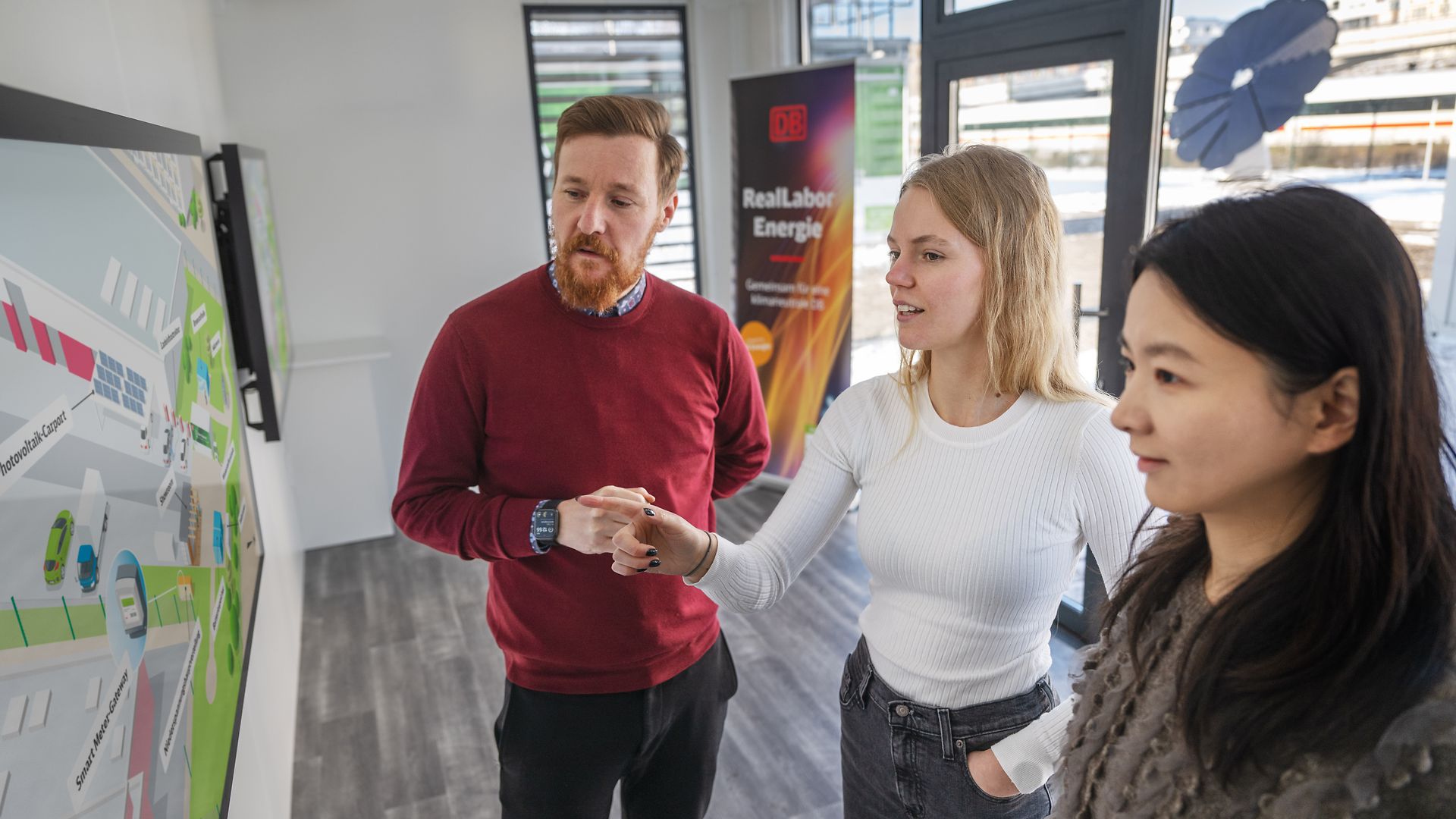 Three people stand in front of a screen in the show room of DB Reallabor Energie.