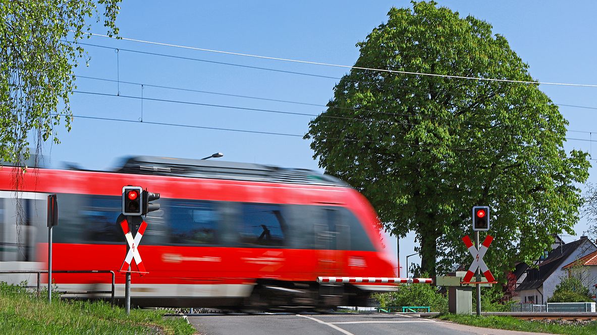 A red train crosses a level crossing in summer, the barriers are closed.