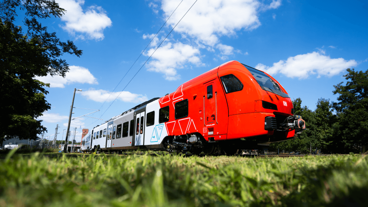 A battery train travels through a green landscape.