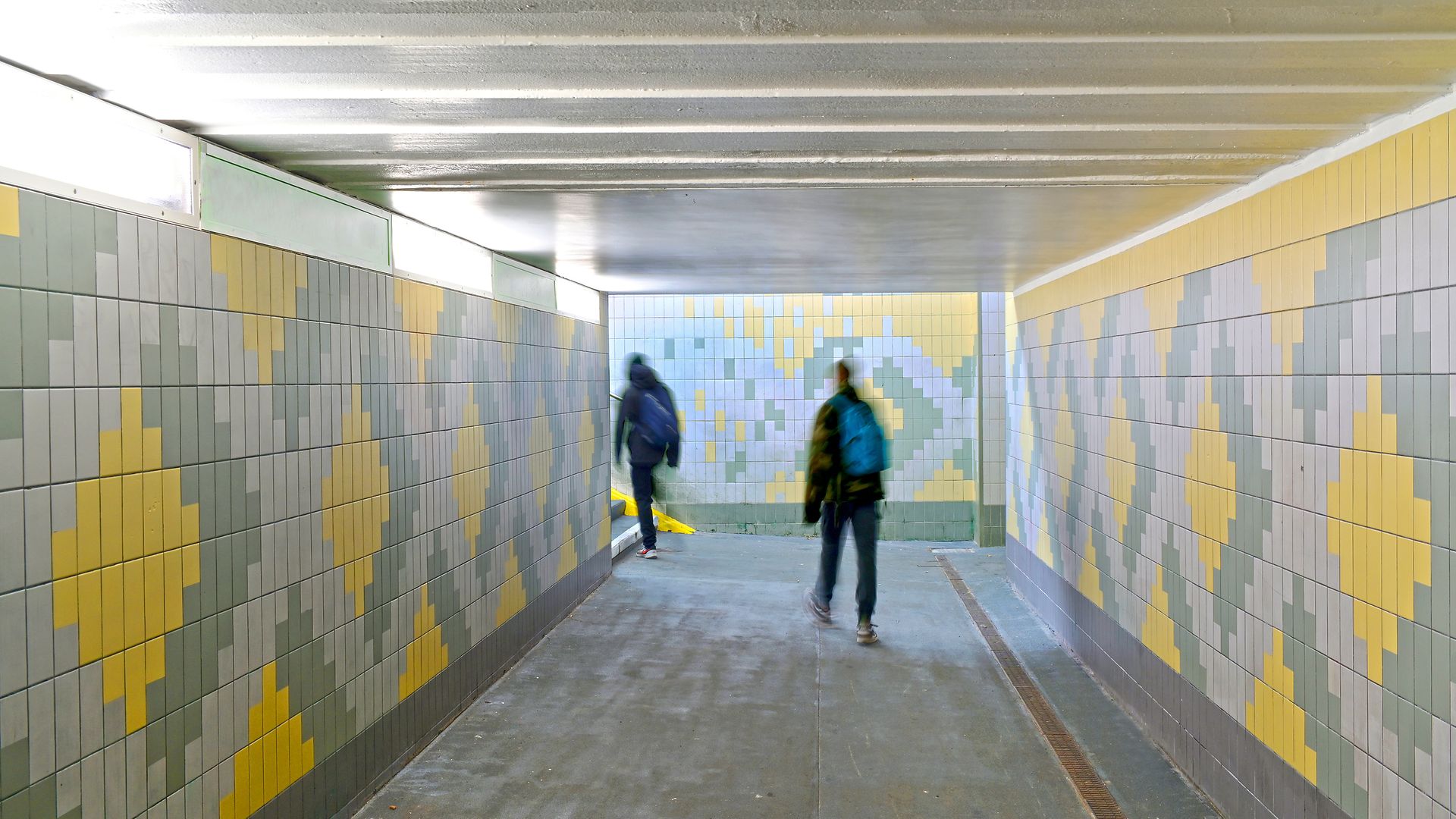 An underpass with yellow and gray tile patterns, two people walking through it.
