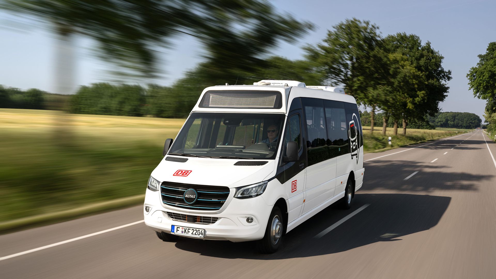 Eine weißer Minibus mit DB Branding fährt auf einer Landstraße.