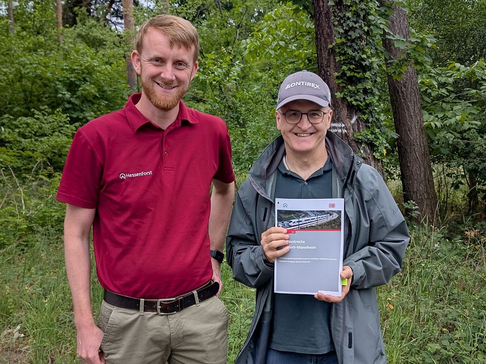 Zwei Personen stehen vor einem Wald und halten eine Kooperationsvereinbarung in der Hand